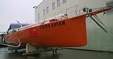 Vu de trois quarts avant, bateau couleur brique à foils blancs posé sur le quai, sans sa quille ni son mât.