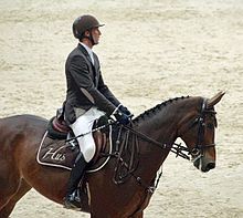 Cavalier de profil en tenue sur sa monture. Sur son tapis de selle est inscrit « Hus » en lettres liées claires.