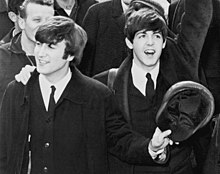 Black-and-white photograph of John Lennon and Paul McCartney in 1964. They are both smiling and waving.