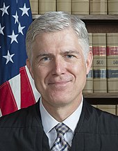 Portrait de Neil Gorsuch dans sa robe de juge devant une bibliothèque et le drapeau américain.