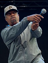 Rapper Chuck D fazendo o movimento de um taco de baseball com o microfone enquanto performa. Ele usa um boné do time de baseball Pittsburgh Pirates.