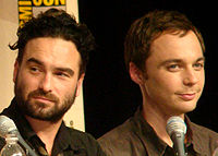 Two caucasian men, one with a beard, sit behind microphones, with a vague symbol in the background.