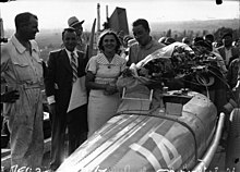 Photo d'un homme en tenue de pilote tenant un bouquet de fleur et félicité par des hommes en costume.