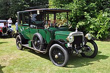 Voiture vert fusain à pare-chocs et toit noirs, roues à rayons, roue de secours sur le marchepied de droite.