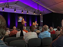 Fiddler Morgan Toney on stage in front of seated crowd along with a guitar player sitting beside Toney.