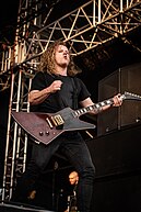 Tyrrell plays guitar with both hands. He has a black T-shirt and black jeans with a tattoo on his right hand.