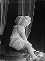 An older baby is sitting on a table. She is wearing a light-colored bonnet with a stiff brim.