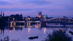 Panorama miasta, na pierwszym planie rzeka, w oddali niskie budynki, dwie wieże kościoła, most