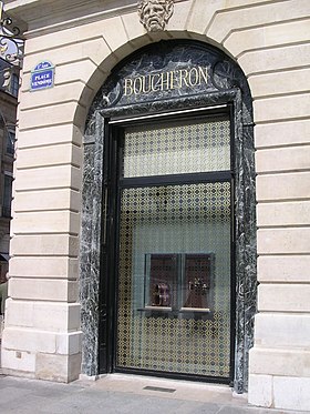 Sur la droite de l'image, le nom Boucheron, gravé en lettres capitales dorées sur un plaquage de marbre sombre, surmontant une vitrine de magasin, et en dessous d'un mascaron décoratif de l'immeuble ; sur la gauche de l'image, une plaque de rue de la place Vendôme.