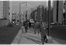 Photo en noir et blanc. Un athlète d'Allemagne de l'Est portant une mallette marche sur le trottoir ; le long de la route, des deux côtés, de nombreux immeubles hauts.