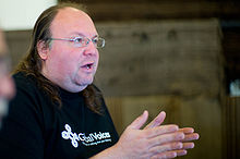 A photograph of a double chin spectacled man with hair slicked back speaks