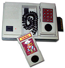 console quadrangulaire grise avec deux manettes en forme de combiné de téléphone. Une cartouche du jeu BurgerTime est posée dessus