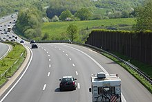 Photographie d'une autoroute prise en surplomb depuis un pont, avec un mur anti-bruit sur la droite et un paysage aux deuxième et troisième plans.