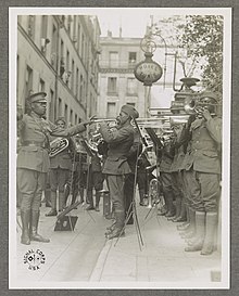 Sur cette photo en noir et blanc, James Reese Europe est au milieu de ses musiciens, qu'il dirige à la baguette. On distingue des trompettes, des trombones, des cors. Les musiciens sont entourés par des bâtiments parisiens, on distingue quelques personnes penchées à leurs fenêtres, et une enseigne indiquant « hôtel tunis ».