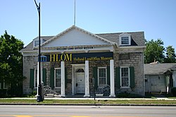 A light brown stone building with a pointed roof, green shutters and white columns on the front. In front of it are a sidewalk, a blue marker with gold writing, and a portion of street. Black letters above the front columns read "Holland Land Office Museum", which is also on a banner across the front.