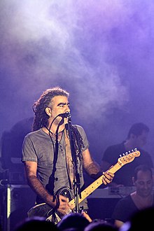 Mosh Ben-Ari playing electric guitar and singing into a microphone on a smoky stage
