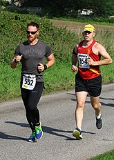 Two persons exercising on a paved road in a park. The weather is sunny.