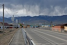 Photographie prise depuis un pont dans le centre du village de Koch-Agatch, composé de maisons. Seulement les pics sont enneigés. Des vaches, au nombre de cinq, circulent sur un côté de la route, avec deux voitures sur la route et un utilitaire qui attend que les vaches passent pour s'engager.