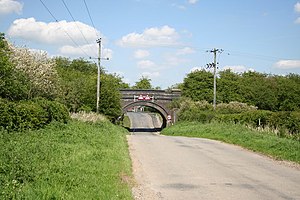 Blue brick single arch bridge carrying former railway over the road into the village. The first houses are just visible through the arch. The road surface has been lowered to pass under the bridge. On either side steep embankments carry the former line, completely overgrown with trees and shrubs