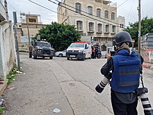 Israeli soldier standing in the street near an intersection with a hospital building in front of the soldier and a military vehicle and ambulance in the intersection.