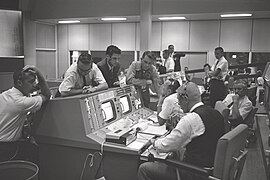 Gene Kranz (devant) et Christopher Kraft (derrière) à leurs consoles du centre de contrôle de mission avec notamment See (en combinaison) le 21 août 1965.