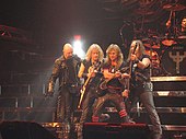 Four men standing next to one another on a stage, three of which are holding guitars. All four men are wearing black clothing, and some of the articles of clothing are studded.