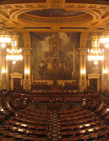 A view of a large, ornately-decorated room with several rows of curved desks, arranged in a semicircle. A large mural is visible on the wall at the far end of the room.