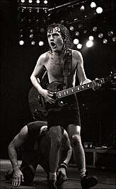 This photo is in black and white. Angus, aged 27, is shown playing guitar with both hands. He is seen shirtless, with shorts, and the socks-shoes combo. Brian Johnson, aged 34, is shown behind Angus, with his hands and knees on the ground. He is seen wearing an Irish hat, a tank top, jeans and shoes. Both are shown in right profile.