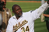 A bald black male baseball player in his fifties pitches a baseball. He is wearing a white uniform with the word "Athletics" across it, and the number 34 below the lettering.