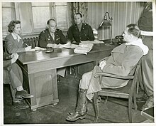 B&W photo of the US Army colonels sitting behind a wooden desk. Rudolf Hess sits in a chair obliquely facing them, right leg crossed over left, fingers interlaced in his lap.