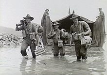 Black and white photo of three men wearing military uniforms and carrying guns and bags wading through water in front of a boat