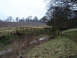 A small wooden footbridge, over a narrow river, connecting two fields