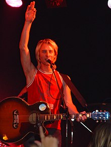 A white man in his mid-thirties stands on a stage. He is reasonably tall, and has light brown hair with blonde highlights. He is holding a microphone in front of his mouth, and has a guitar in his hand. He is wearing jeans and a red top.