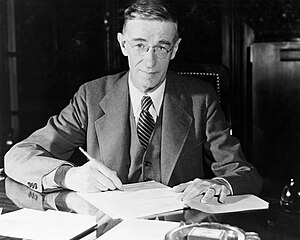 A man in a suit poses sitting at a desk, pen in hand. Papers and an ash tray are on the desk.