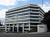 A nine-storey office building with prominent white-panelled storey bands between blue-tinted windows. In the foreground is a white block with the American Express logo.