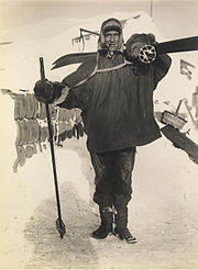 Man, standing, wearing a smock, heavy trousers and boots. He has a ski stick in his right hand, a pair of skis strapped on his back, and is carrying a rounded bundle on his shoulder. Behind him on the ground is assorted polar equipment.