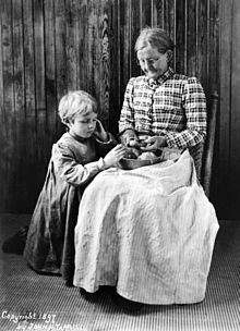 Portrait en noir et blanc d’une femme âgée et d’un enfant posant devant un lambris de bois : la femme, assise, épluche au couteau une pomme de terre au-dessus d’un plat posé sur ses genoux tandis que l’enfant, à genoux sur sa droite, examine un tubercule du récipient. Le maintien de la femme est guindé mais le visage incliné et attentif est légèrement souriant ; elle porte des lunettes rondes et les cheveux gris tirés en chignon, un corsage à carreaux et un ample tablier clair, ligné, qui descend jusqu’au sol. L’enfant aux cheveux courts et blonds, semble très sérieux ; il porte un long tablier à carreaux vichy dont les longues manches sont resserrées aux poignets.