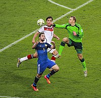 Neuer, in a Germany goalkeeper jersey, leaps into the air to punch the ball away from Argentina player Gonzalo Higuaín while Germany player Mats Hummels covers Neuer