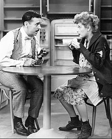 Two characters sit at the table and chat with expressive faces, a common scene in sitcoms