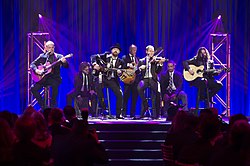 A group of seven men in dark suits playing various musical instruments on a stage in front of an audience