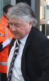 A grey-haired man in a black suit walks out of a building.