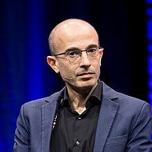 A slender middle-aged bald man with glasses rests his chin on his hand. He is dressed up.