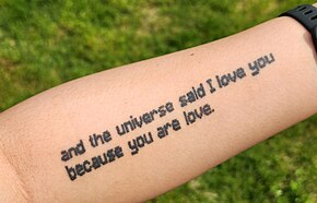 A pale white arm, tattooed with the quote "and the universe said I love you because you are love", all lowercase and in a Minecraft-like font.