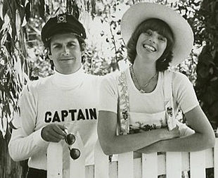 A man wearing a nautical hat and a sweater with "Captain" on it, and a woman wearing a broad-brimmed hat and dungarees leaning on a picket fence