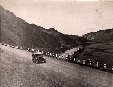 Photographie d'une route en gravier bordant une falaise, avec une rivière qui coule en contre-bas dans la vallée. Au centre de la photographie, une voiture des années 1930 circule de manière confortable. La route est lisse, recouverte de gravier.