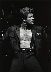 A dark-haired young man wearing a dark jacket open to reveal only suspenders holding up his pants
