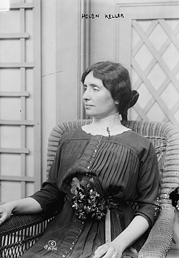 Photographie en noir et blanc montrant Helen Keller assise dans un fauteuil d'osier sur le pas d'une porte. Elle porte une robe ajustée sombre, et elle a des cheveux sombres et ondulés rassemblés en chignon. Une inscription à la plume indique son nom en haut de la photo.
