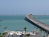 A long bridge goes across a large body of light blue water oriented left to right, with the skyline of a city on the horizon.