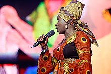 Angélique Kidjo de profil, portant un pagne wax de couleur dominante orange, tenant un micro sans fil dans sa main droite.