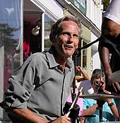 A man wearing a dress shirt, sitting behind a drum kit; spectators can be seen in the background.